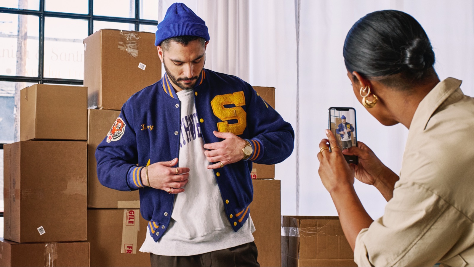 Woman taking a photo on her iPhone of a man wearing a blue jacket standing in front of packing boxes.