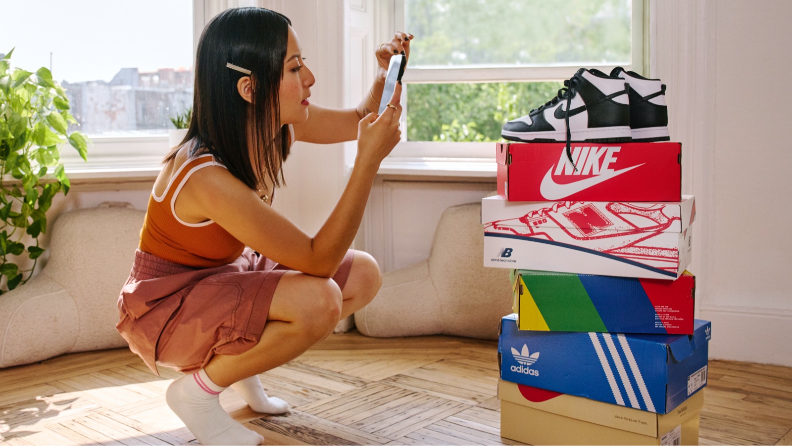 Woman crouching down, taking a picture on her phone of sneakers on top of a stack of sneaker boxes.